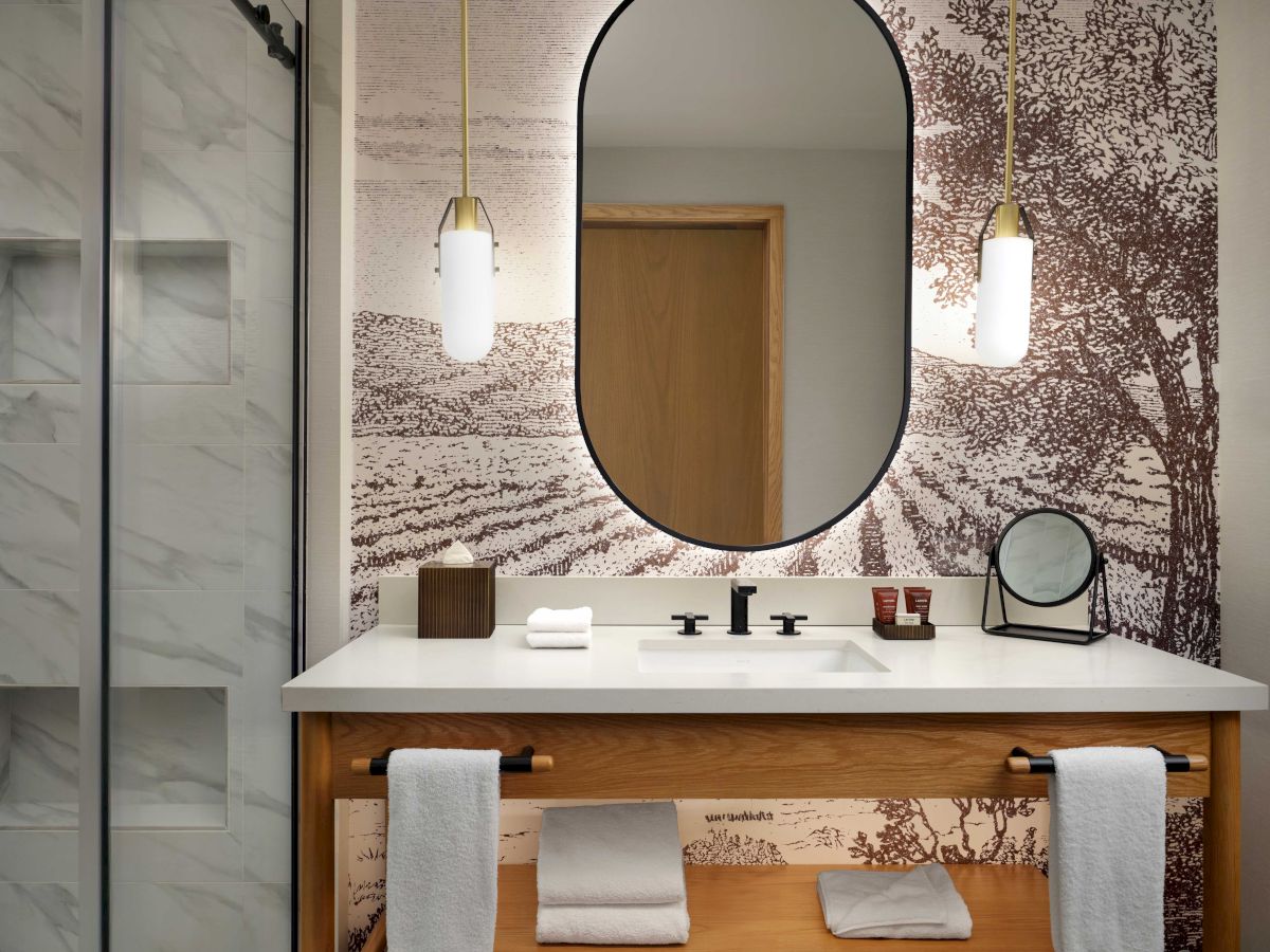 A bathroom with an oval mirror, pendant lights, wooden vanity, towels, and a scenic wall design.