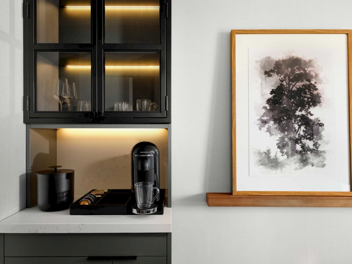 A countertop with a coffee maker, cups, and a cabinet with glasses. A framed abstract art piece is on the wall to the right.