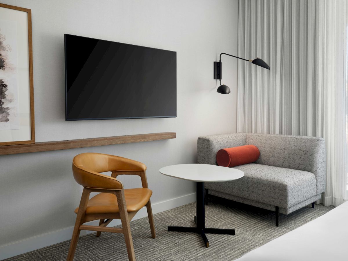 The image shows a modern hotel room with a wall-mounted TV, chair, small table, sofa, and a framed artwork on the wall.