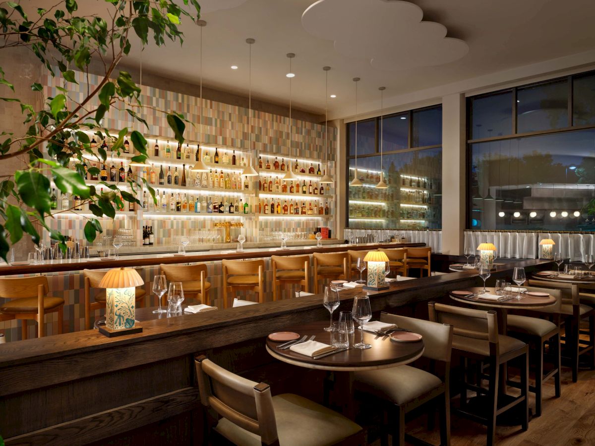 Cozy, modern restaurant with warm lighting, wooden bar and seating, wine shelves along back wall, large windows, and greenery accents. ending with a period.