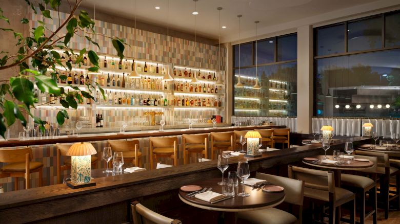 Cozy, modern restaurant with warm lighting, wooden bar and seating, wine shelves along back wall, large windows, and greenery accents. ending with a period.
