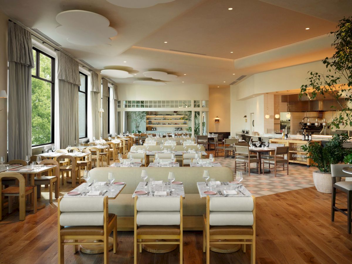 A bright, airy dining room with long tables set for a formal meal, soft lighting, wooden floors, and large windows with plants and modern seating.