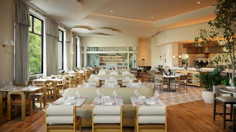 A bright, airy dining room with long tables set for a formal meal, soft lighting, wooden floors, and large windows with plants and modern seating.