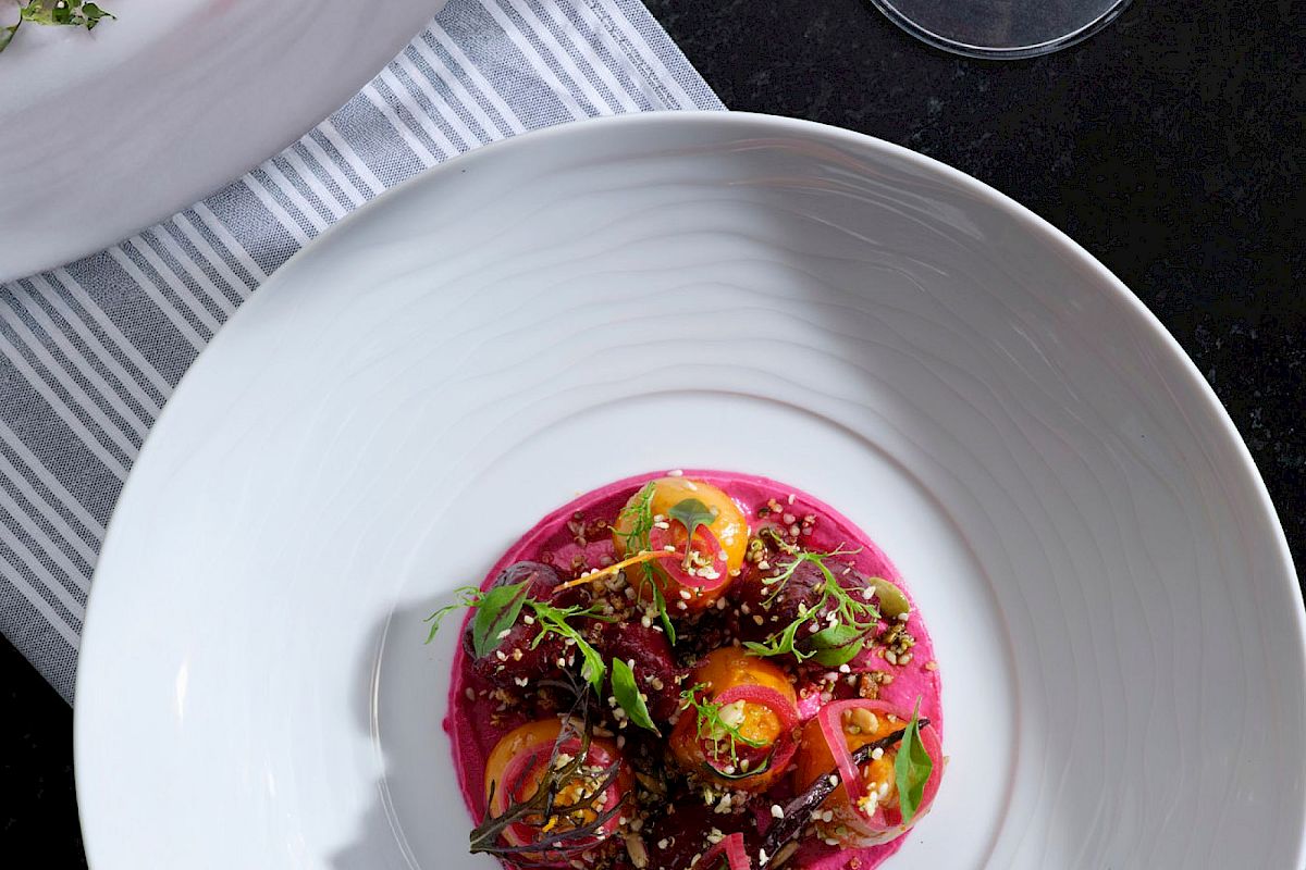 A gourmet dish with colorful vegetables and greens on a pink puree, next to a glass of red wine on a striped cloth.