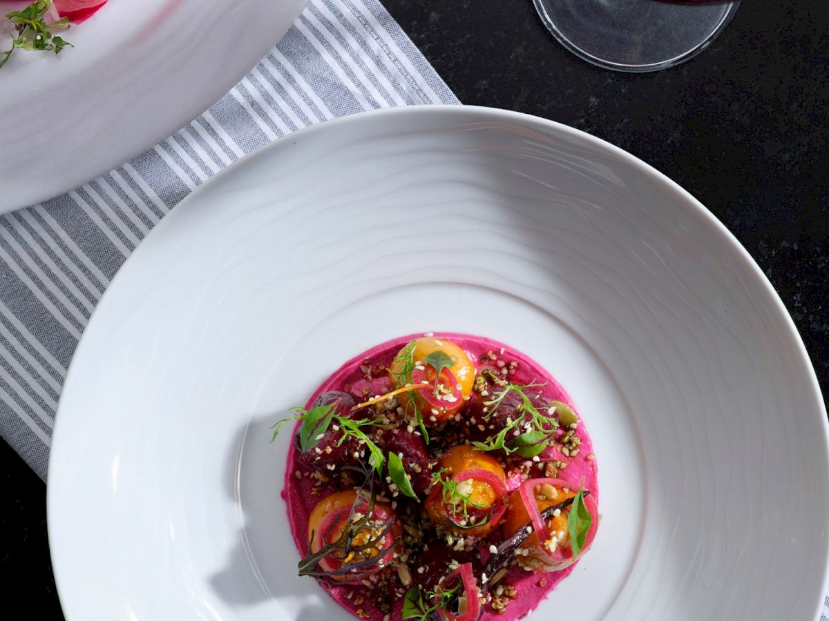 A plated dish with vibrant pink beet puree topped with greens and herbs, served in a white bowl on a striped placemat.