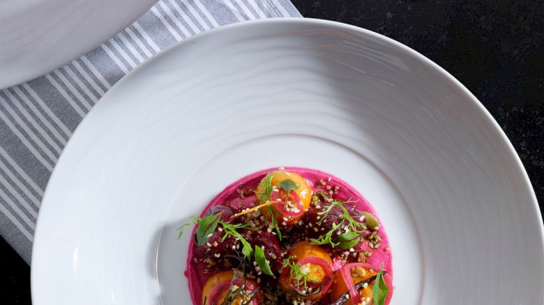 A gourmet dish with colorful vegetables and greens on a pink puree, next to a glass of red wine on a striped cloth.
