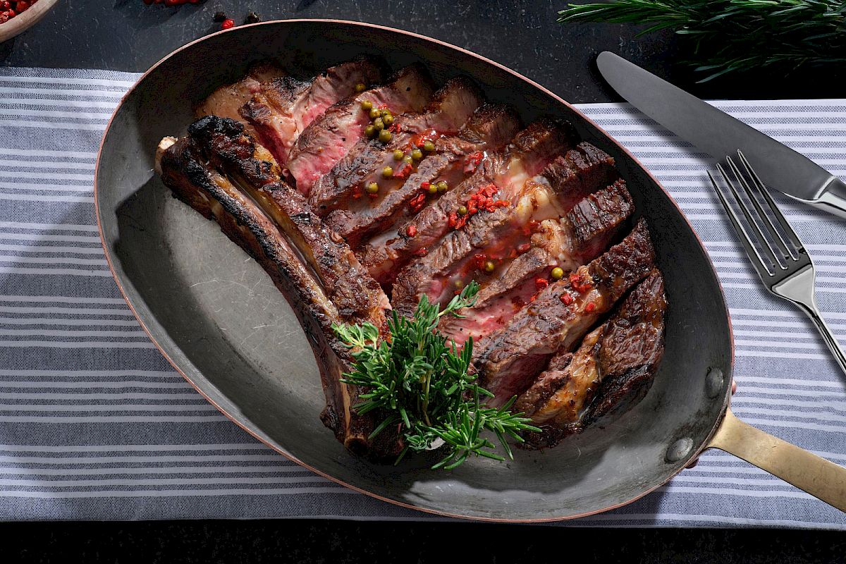 A sliced, grilled tomahawk steak garnished with herbs and peppercorns, served on a pan with a striped cloth, knife, and fork beside it.