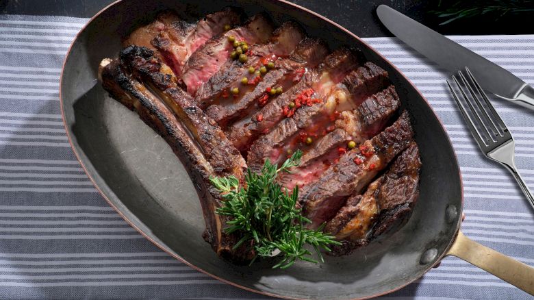 A sliced, grilled tomahawk steak garnished with herbs and peppercorns, served on a pan with a striped cloth, knife, and fork beside it.