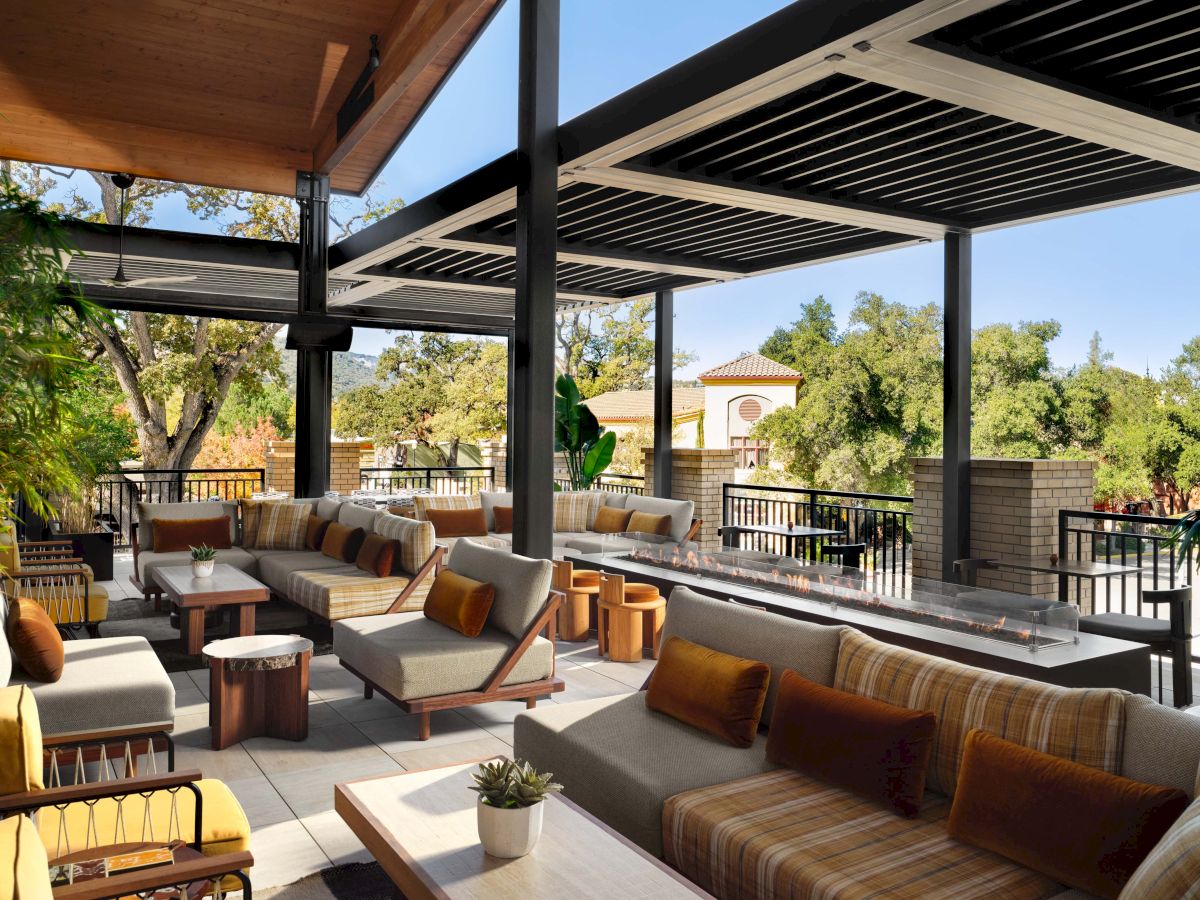 A stylish outdoor lounge with striped couches, wooden tables, and a shaded patio overlooking trees under a sunlit, modern veranda.