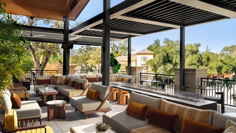 A stylish outdoor lounge with striped couches, wooden tables, and a shaded patio overlooking trees under a sunlit, modern veranda.
