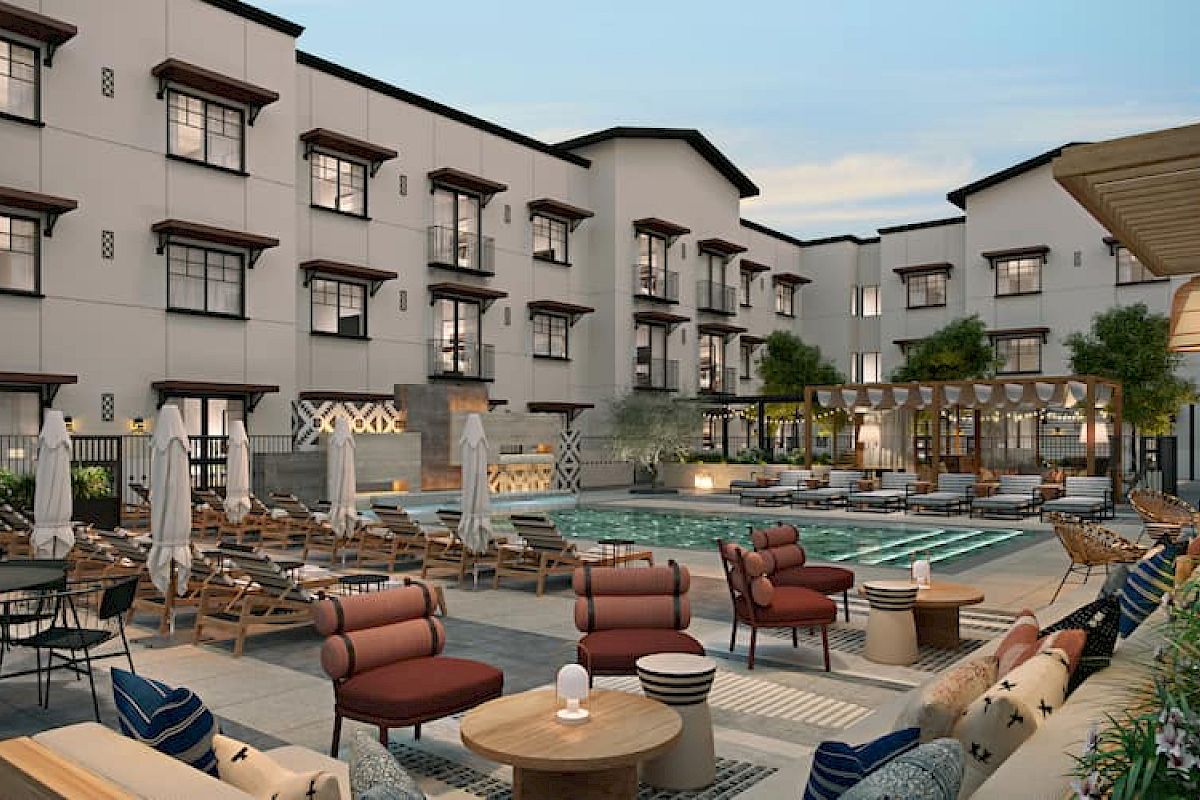 This image shows a hotel courtyard with a swimming pool, surrounded by seating, umbrellas, and a building with multiple windows.