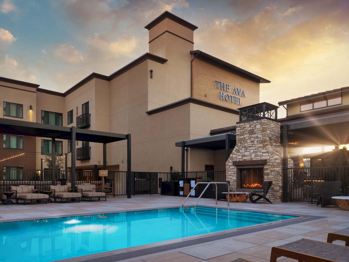A hotel’s outdoor pool area at sunset, with a beige building, lounge chairs, a stone fireplace, and a bright blue pool reflecting the warm sky.