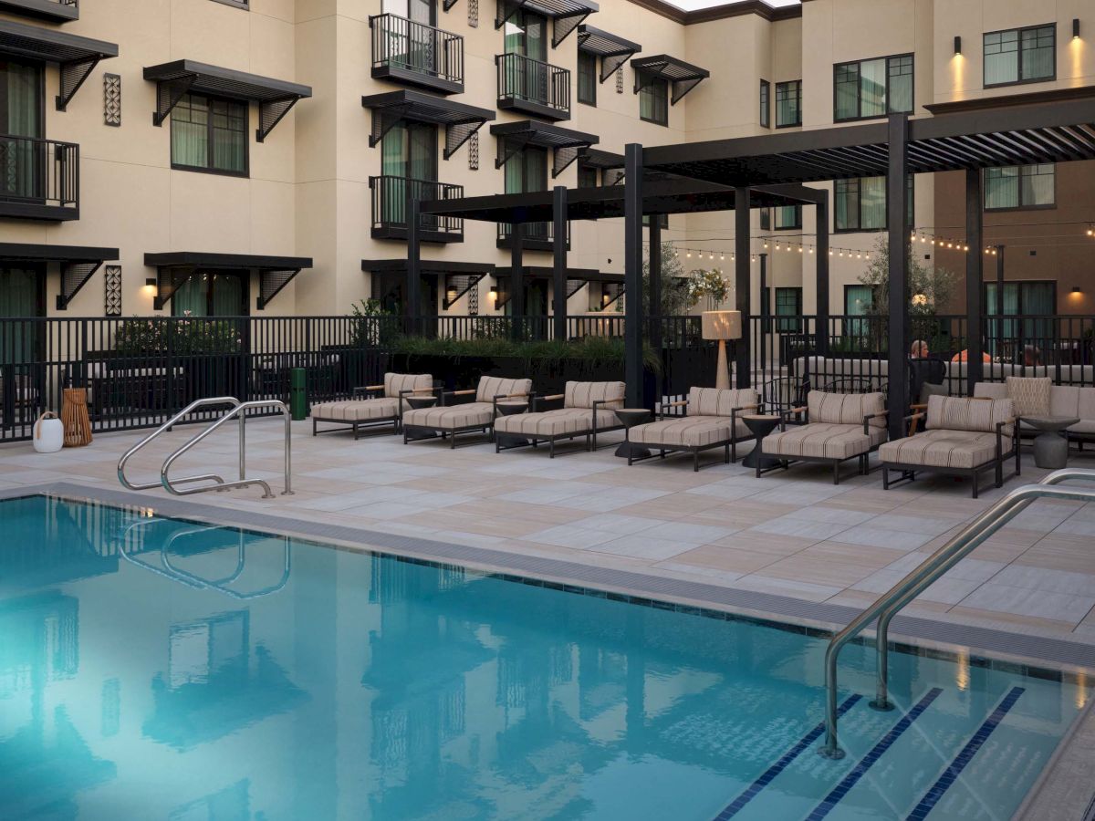 A hotel courtyard with a blue swimming pool, sun loungers, and a modern beige building surrounding the deck, viewed at sunset.