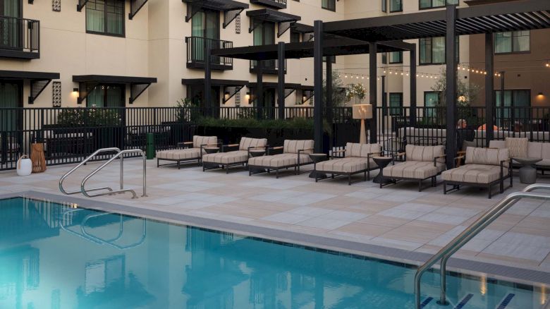A hotel courtyard with a blue swimming pool, sun loungers, and a modern beige building surrounding the deck, viewed at sunset.