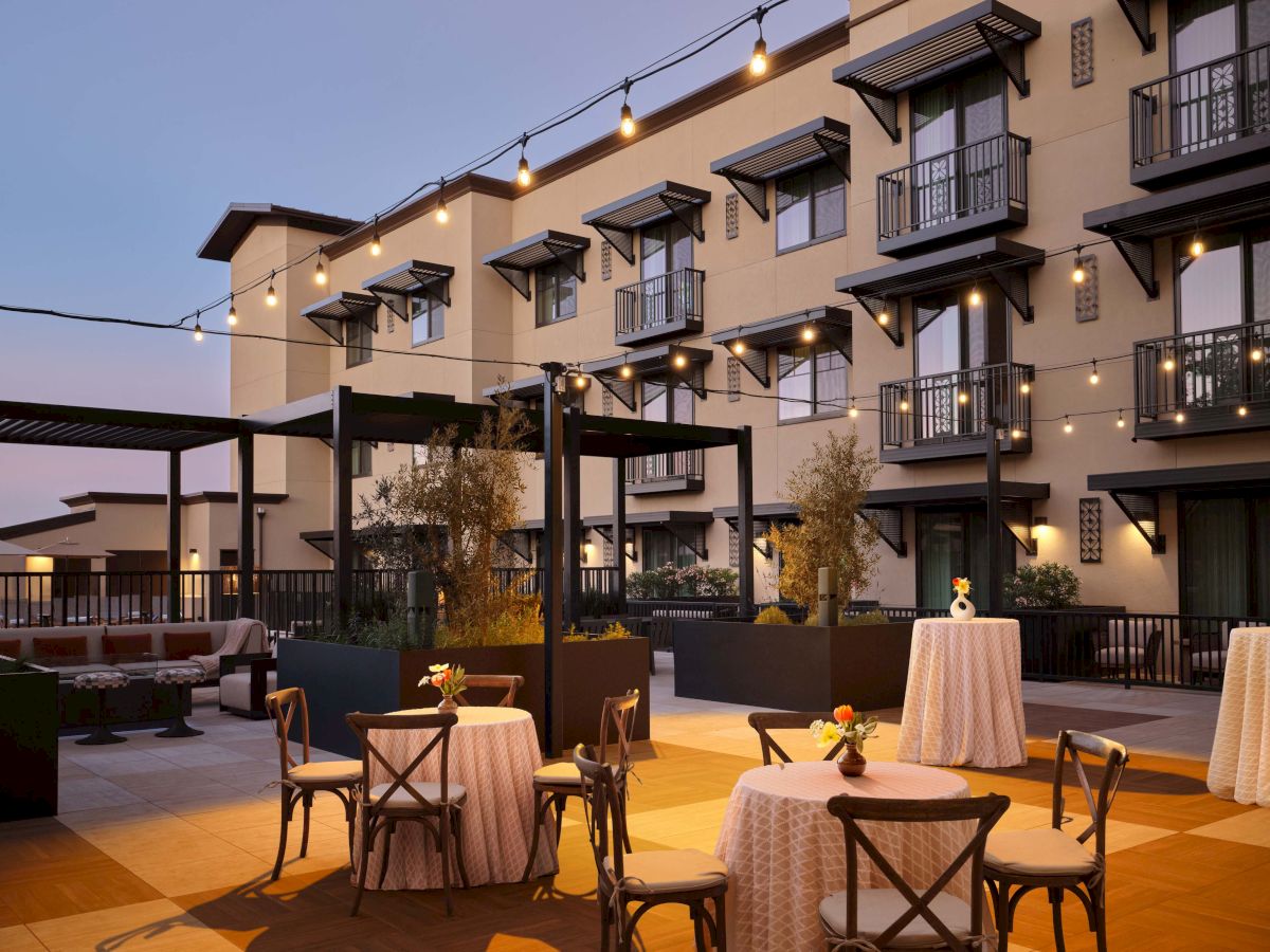 Outdoor courtyard at dusk with string lights, small round tables, elegant chairs, and a multi-story hotel building in the background.