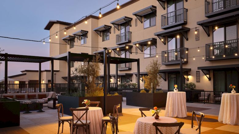 Outdoor courtyard at dusk with string lights, small round tables, elegant chairs, and a multi-story hotel building in the background.