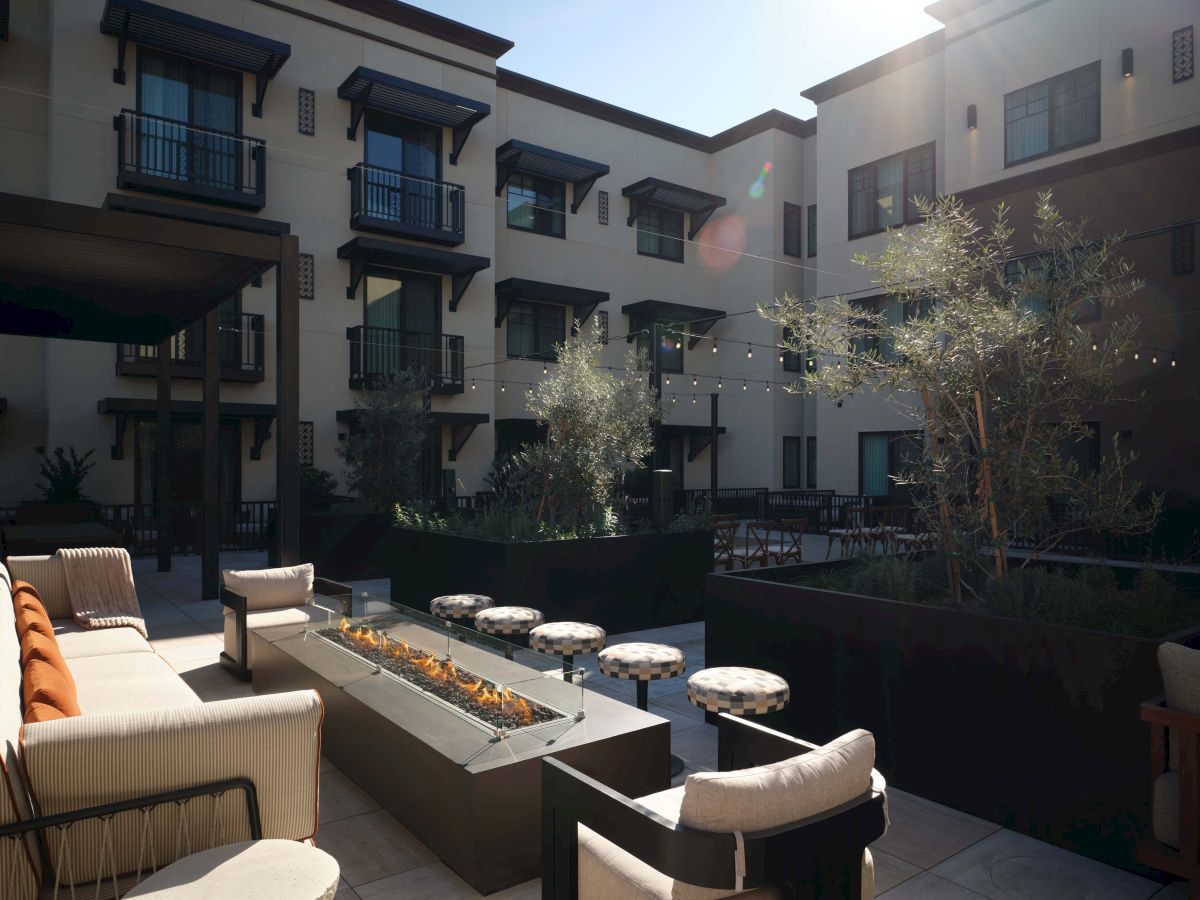 A modern hotel courtyard with outdoor seating, string lights, planters, and a sunlit, three-story building in the background.