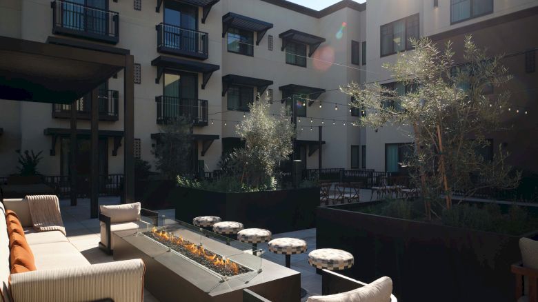 A modern hotel courtyard with outdoor seating, string lights, planters, and a sunlit, three-story building in the background.