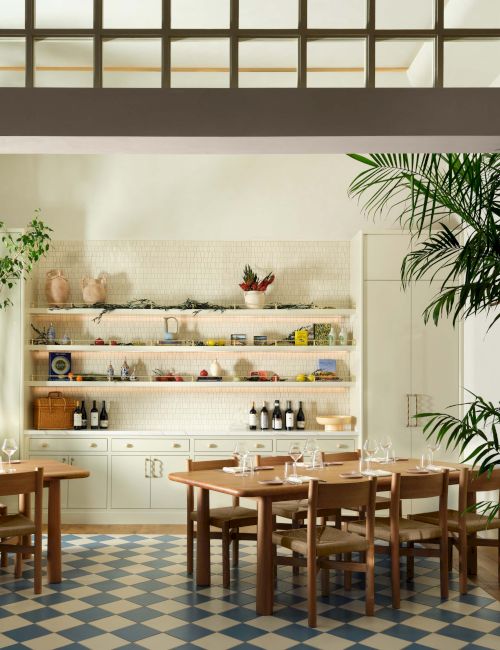 A bright dining area with wooden tables and chairs, a checkered floor, greenery, and a small open kitchen with shelves.