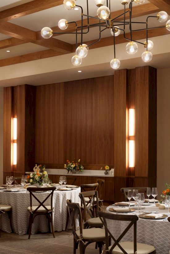 A stylish dining room with wooden paneling, soft lighting, and round tables set for a formal meal, creating an warm, elegant ambiance.