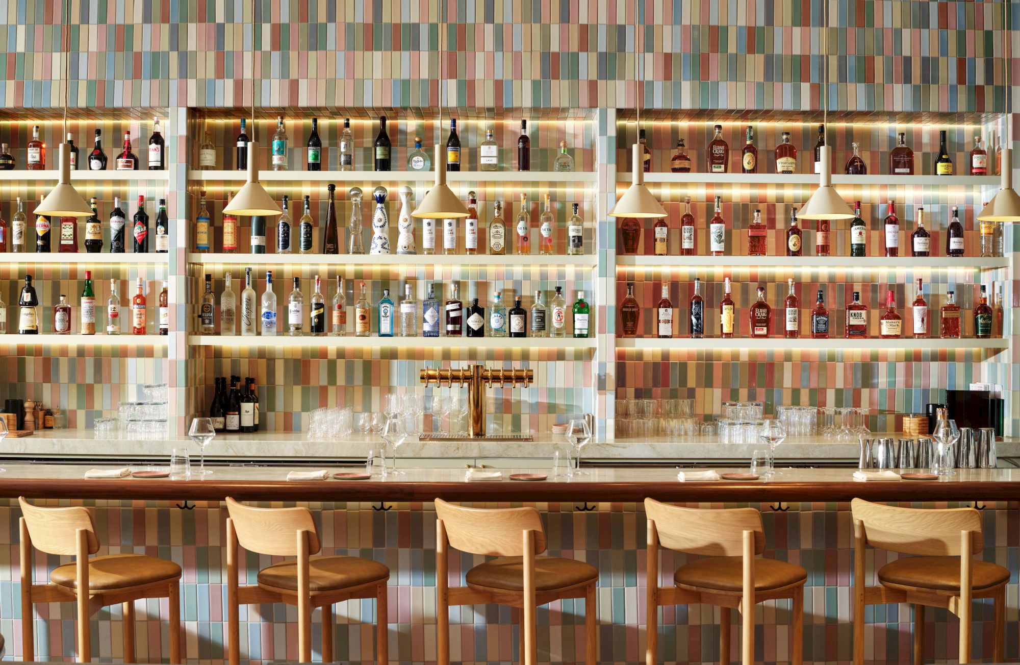 A stylish bar lined with wooden stools, colorful tile backdrop, and shelves full of bottles, inviting cocktails and casual conversation.