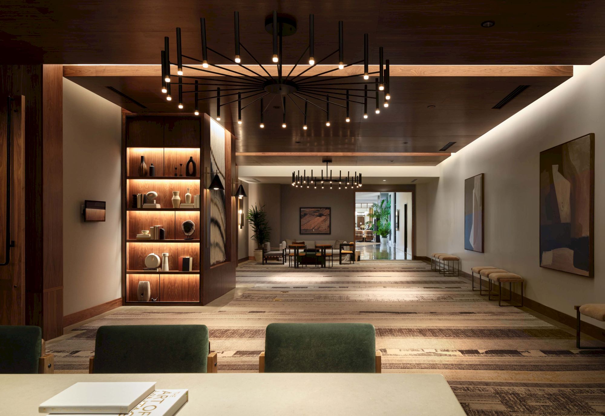 A stylish, modern lobby with wooden beams, warm lighting, a bookshelf display, long bench seating, and a coffee table in the foreground, open to a bright, inviting sitting area.