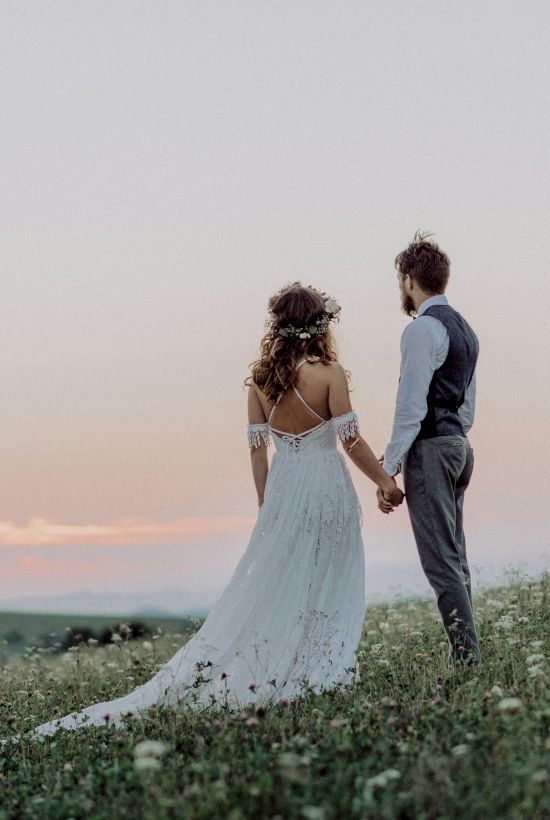 A couple in formal attire stands on a grassy hill, holding hands and gazing at a scenic, pastel sunset in the distance.