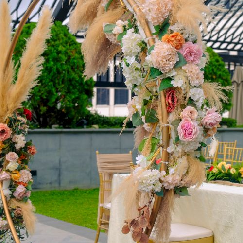 The image shows a decorated outdoor event space with floral arrangements and pampas grass, alongside chairs and a table with a cloth.