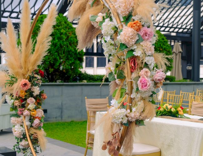 The image shows a decorated outdoor event space with floral arrangements and pampas grass, alongside chairs and a table with a cloth.