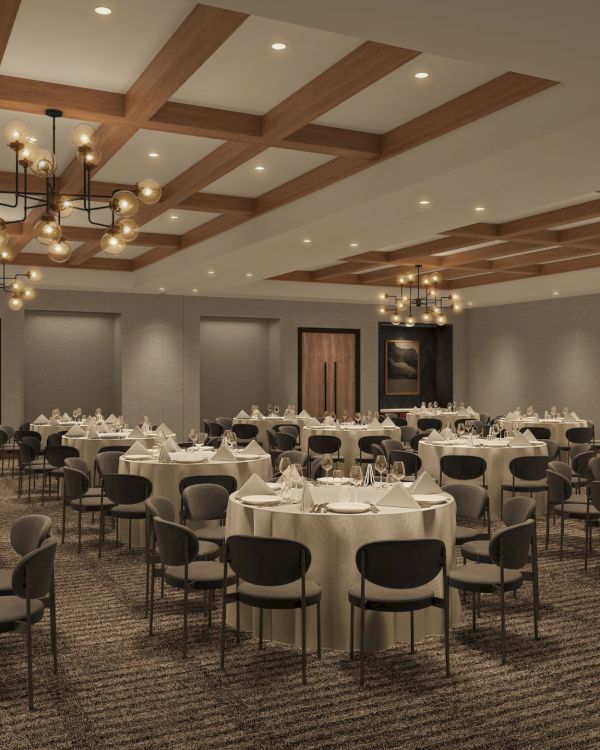 A modern banquet hall with round tables set for dining, elegant chandeliers, wooden paneling, and neutral decor.