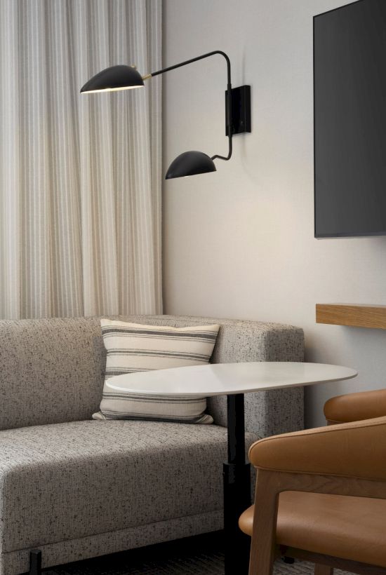 A cozy seating area with a gray sofa, striped pillow, small round table, leather chair, wall-mounted lamp, TV, and sheer curtains.