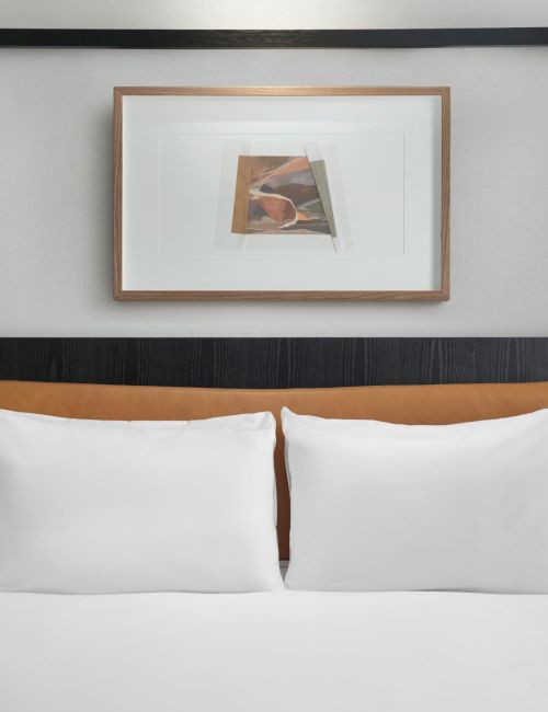 A neatly made bed with white pillows, a padded brown headboard, flanked by lamps, and a framed artwork above.