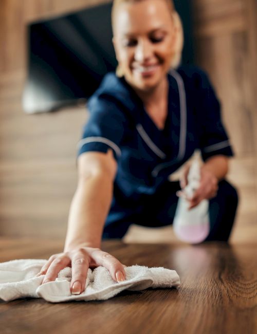 A person in uniform is wiping a wooden table with a cloth, holding a spray bottle, and smiling while cleaning a room.