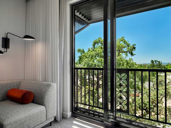 A bright hotel room with a gray sofa and orange bolster, a wall lamp, sheer curtains, and a balcony door opening onto a green landscape with blue sky.