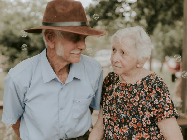 An elderly couple outdoors, the man in a hat and blue shirt, and the woman in a floral dress, standing closely together and smiling.