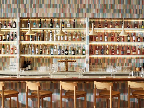 A stylish, well-lit bar with a colorful tile backdrop and a long counter lined with wooden stools.