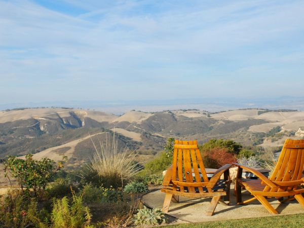 Two wooden chairs overlook a scenic landscape of rolling hills under a clear blue sky.