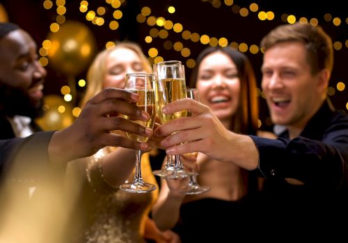 A group of people celebrating, clinking glasses, with festive lights in the background.