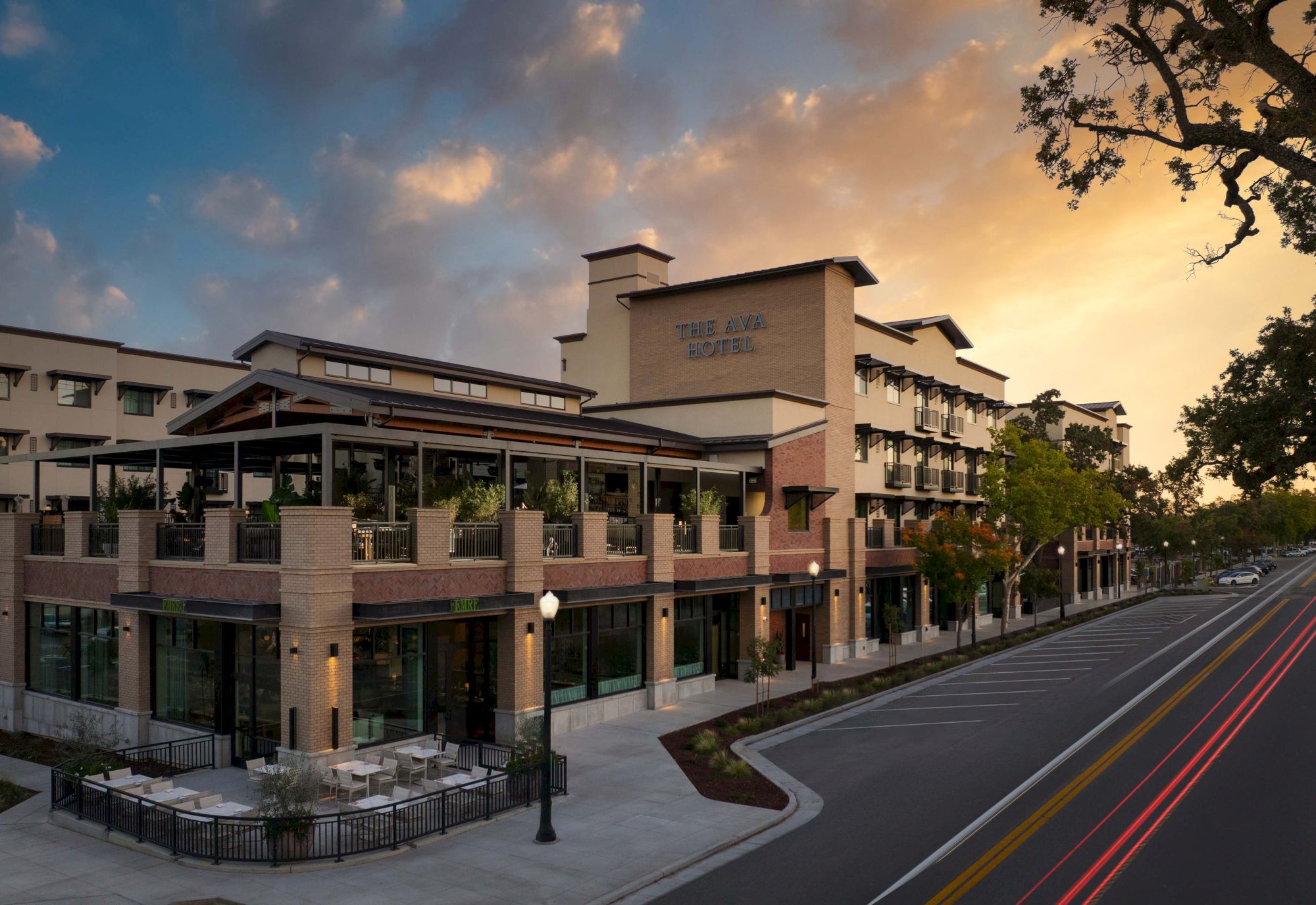 A modern shopping and dining complex with a corner cafe, brick accents, and a tree-lined street at sunset.