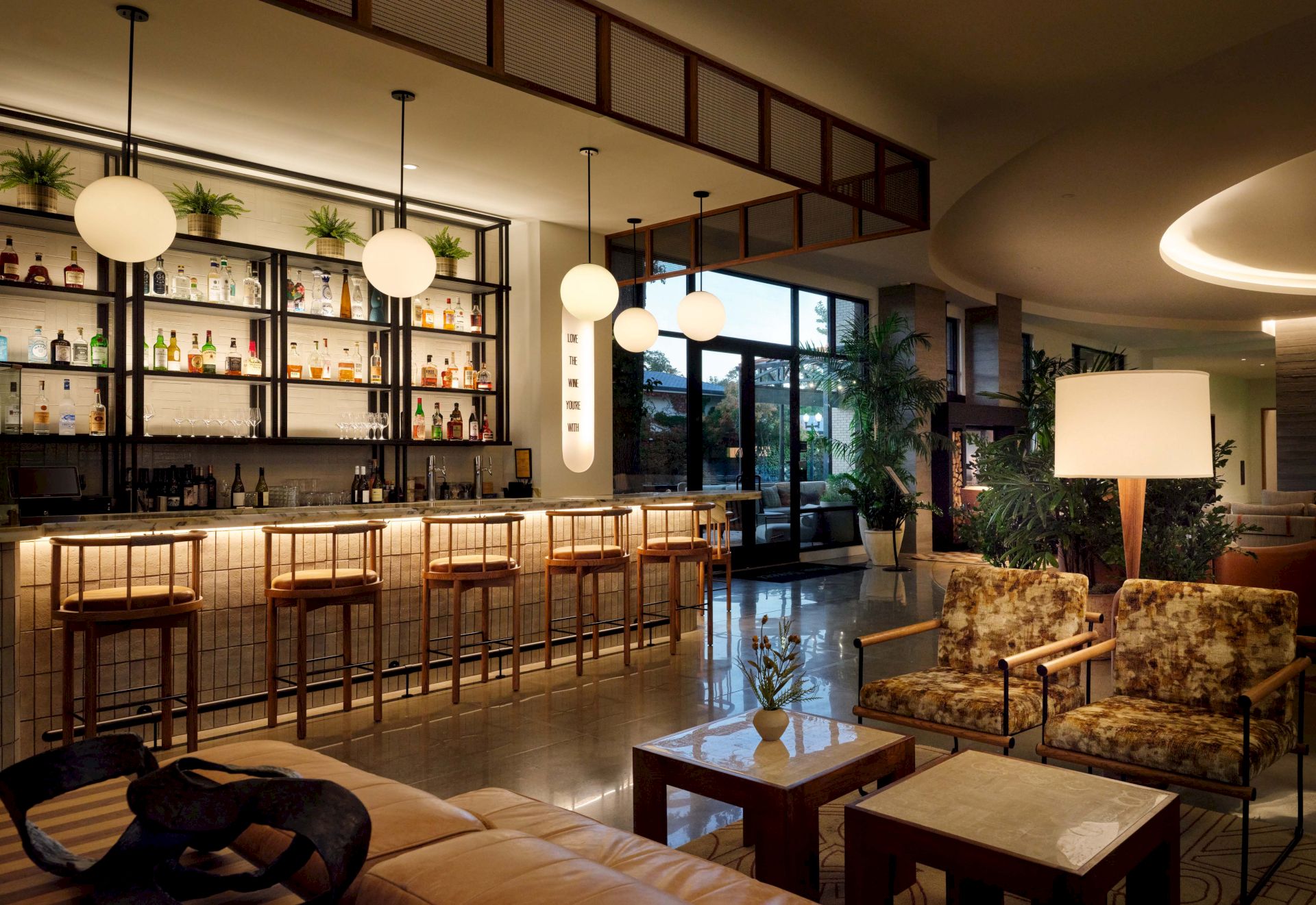 A stylish hotel lounge bar with a long wooden bar, circular pendant lights, bar stools, and a cozy seating area with plants and lamps.