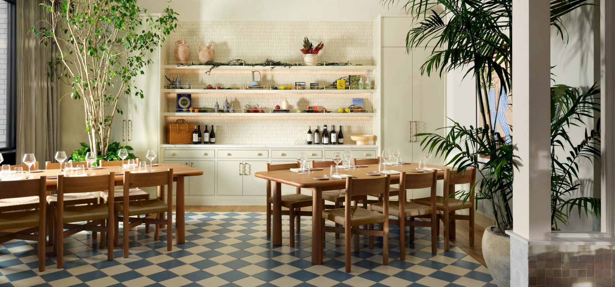 A bright, airy dining area with wooden tables and chairs, a tiled blue-and-white floor, greenery, and a white open shelf kitchen setup in the back, end.
