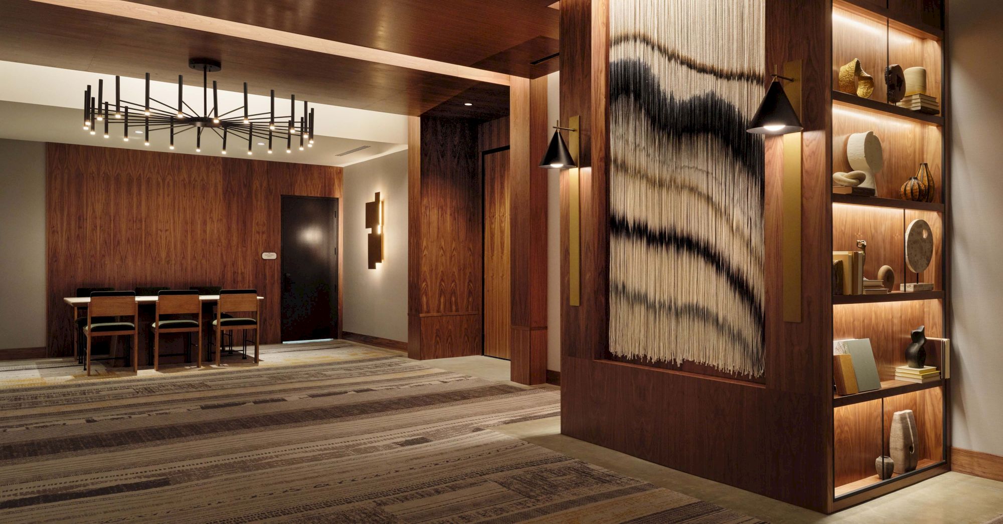A stylish hotel lobby or lounge with dark wood paneling, a dining table area in the back, ambient lighting, and a decorative shelf wall with art and shelves.
