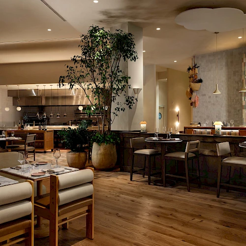 A stylish restaurant interior with wooden floors, comfortable seating, plants, warm lighting, and a bar area in the background.