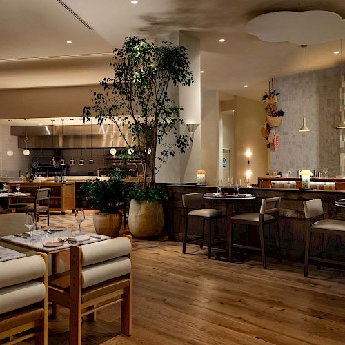 A stylish restaurant interior with wooden floors, comfortable seating, plants, warm lighting, and a bar area in the background.