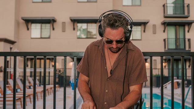 A DJ wearing sunglasses and a brown shirt spins records at a poolside setup, with a patterned table, gear, and a hotel-style building in the background.