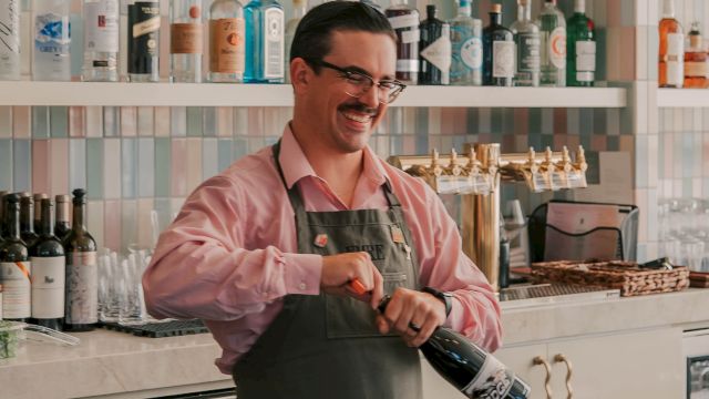 A smiling bartender in a pink shirt and apron pops a wine cork behind a well-stocked bar with shelves of bottles; cheerful, lively scene.