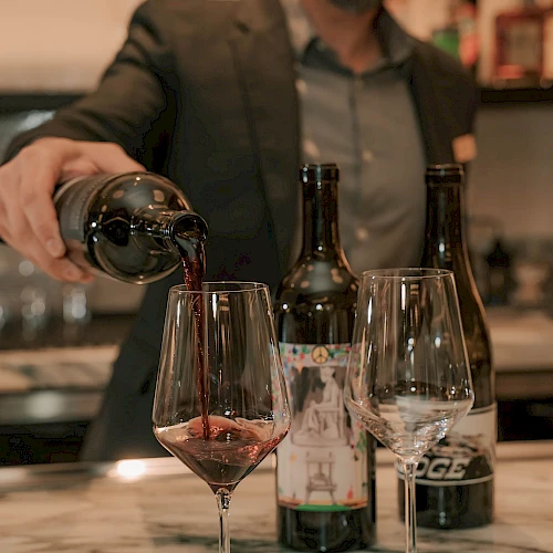 A bartender pours red wine into a glass at a marble bar, with two bottles and two empty glasses nearby, ending this sentence.