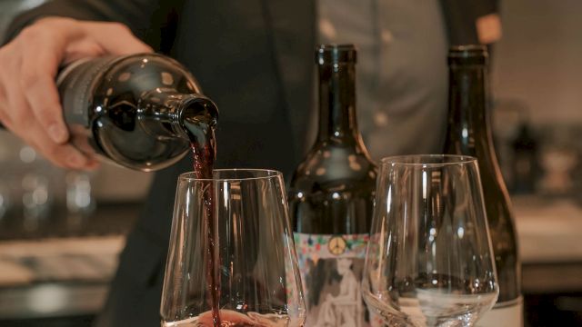 A bartender pours red wine into a glass at a marble bar, with two bottles and two empty glasses nearby, ending this sentence.