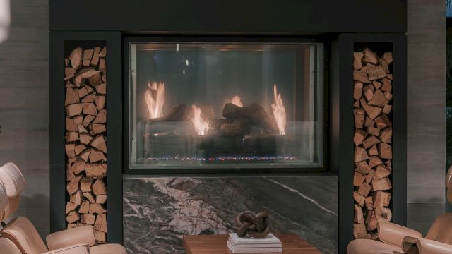 A modern living room with a lit stone fireplace centered between built-in log racks, flanked by beige leather chairs and a wooden coffee table, cozy mood.