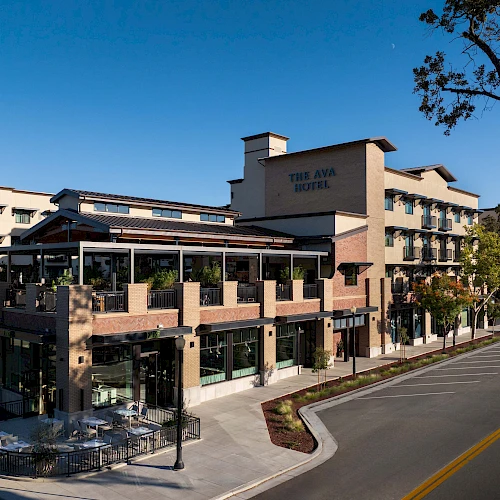 A modern hotel or mixed-use building with brick accents, patio seating, and a street lined with trees under a clear blue sky.
